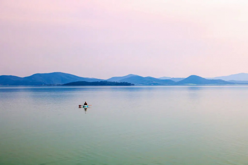   Kuva on otettu Trasimenojärvellä kauniina kevätpäivänä ja siinä näkyy yksinäinen kanootti keskellä järveä ja sen takana yksi saarista sekä kukkuloita ja vuoria horisontissa.