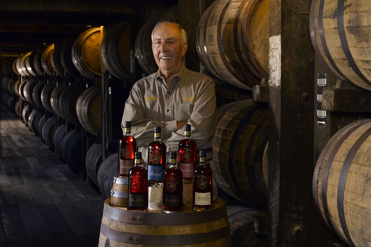 Un hombre detrás de un barril coronado con botellas de whisky, pared de barriles detrás de él.