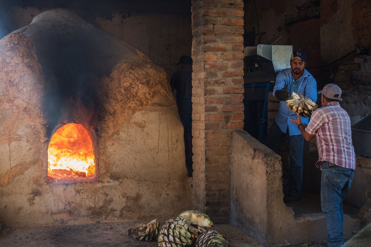 Gran horno de barro con fuego rugiente, dos hombres pasando corazones de agave