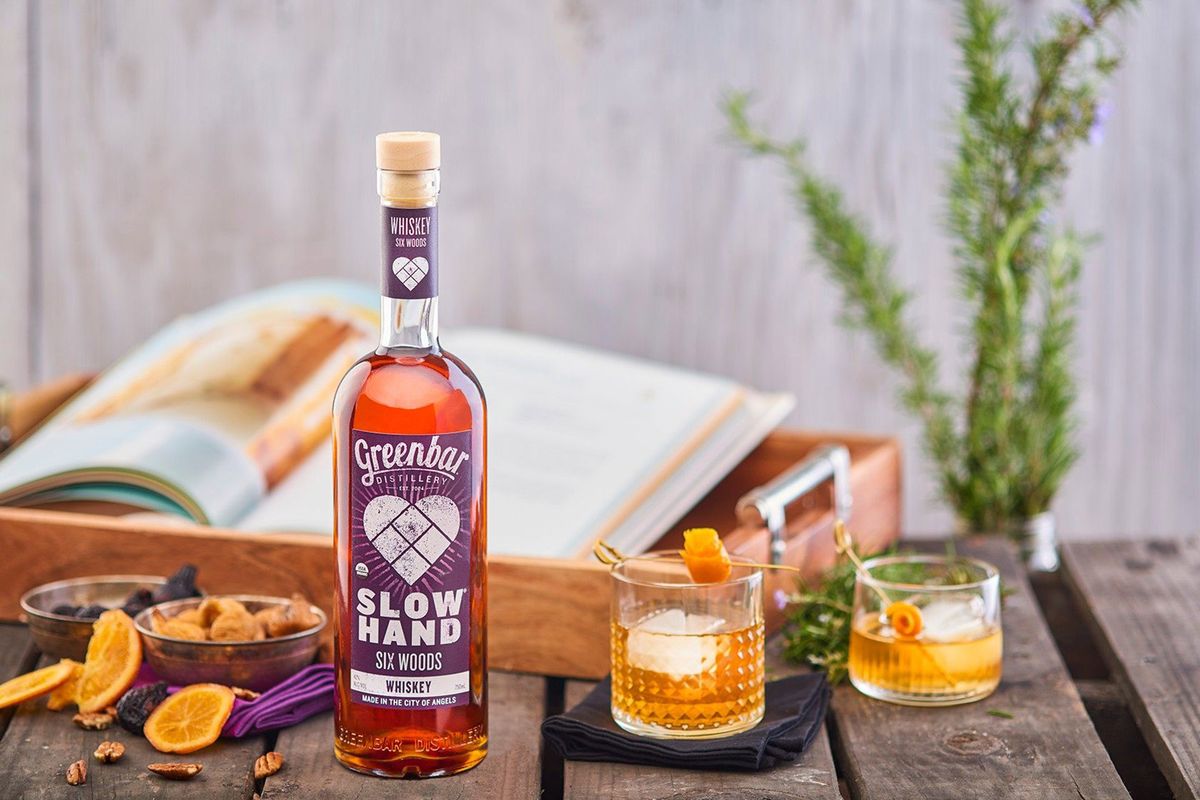 En slank flaske whisky med lilla mærkning, to hælder på klipperne, nødder, frugt og en sløret bog på et træbord