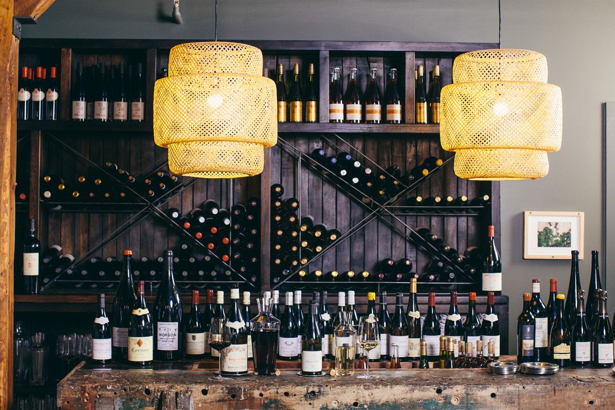 Nhiều chai rượu trên mặt bàn gỗ, tủ đựng rượu trên tường gỗ phía sau
