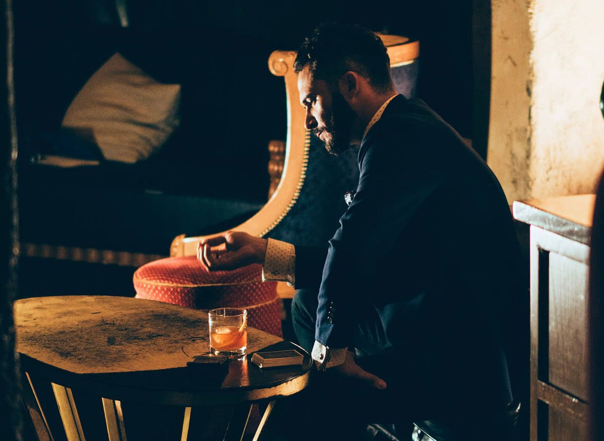 Sitzender Mann, der nachdenklich auf seine Hand, seinen Cocktail und ein Kartenspiel auf dem Tisch vor ihm schaut