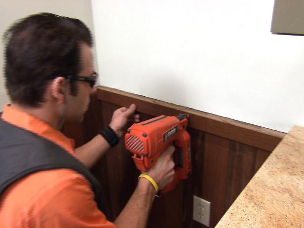 Mennesket bruger en neglepistol til at installere et forklæde omkring wainscoting på væggen.