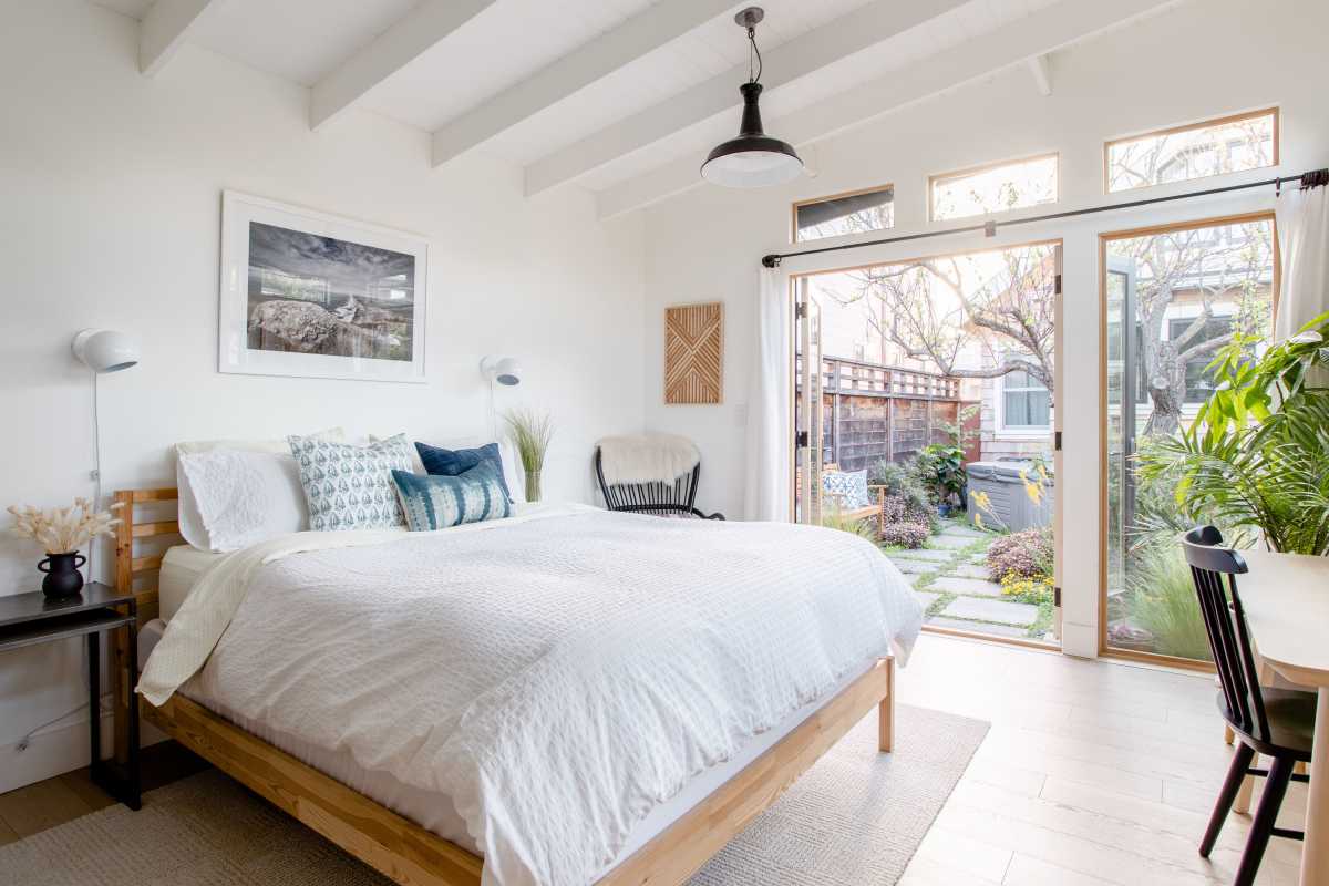 Una cama grande en un espacioso dormitorio blanco, con una puerta corrediza de cristal que da a un patio exterior.