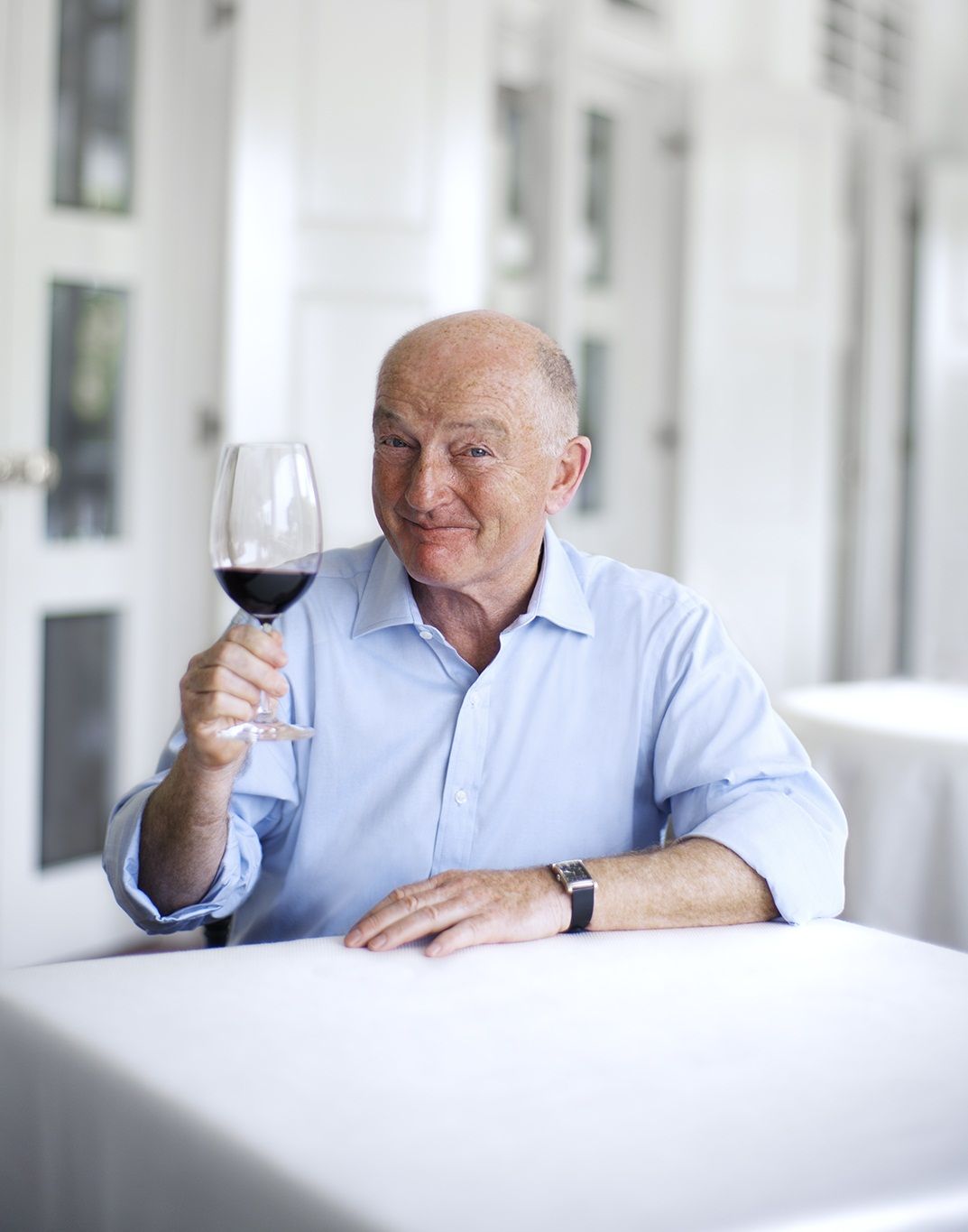 Starejši moški v modri srajci, ki drži kozarec rdečega vina za belo mizo