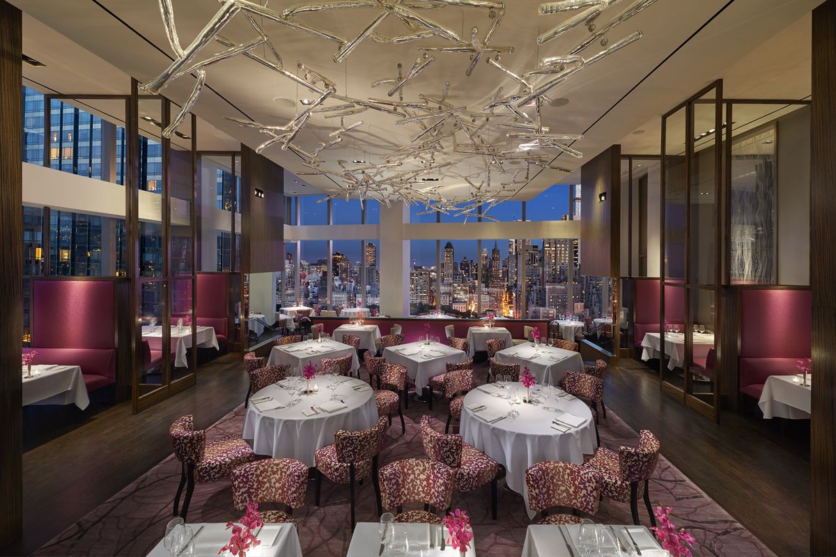Fushia i biała restauracja, wysokie oparcia po obu stronach, biało ubrane stoły pośrodku, dalej, nocna panorama Nowego Jorku