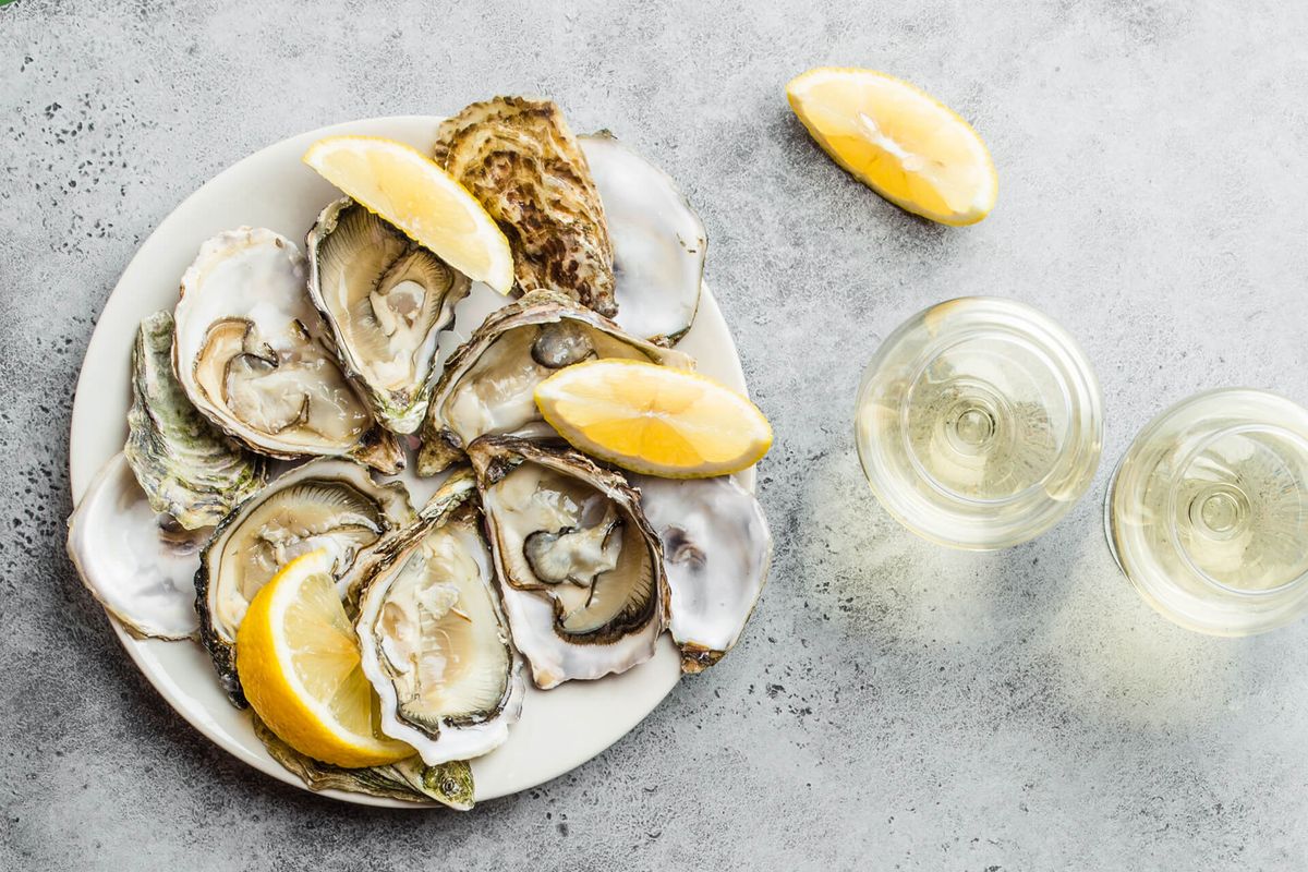 Primer plano de media docena de ostras y conchas frescas abiertas con rodajas de limón en un plato, dos vasos de vino blanco, vista superior, fondo de hormigón rústico gris, espacio para texto