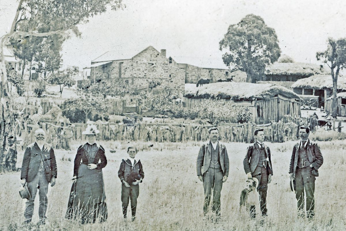 Hombre y mujer de edad avanzada en traje victoriano, tres hombres a la derecha de ellos, gran granja detrás