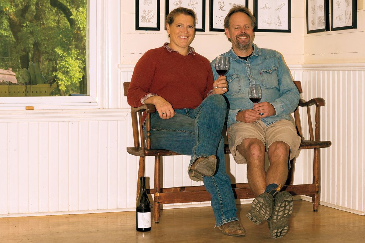 Clare Carver e Brian Marcy sentados em um banco de madeira em uma casa, segurando taças de vinho tinto