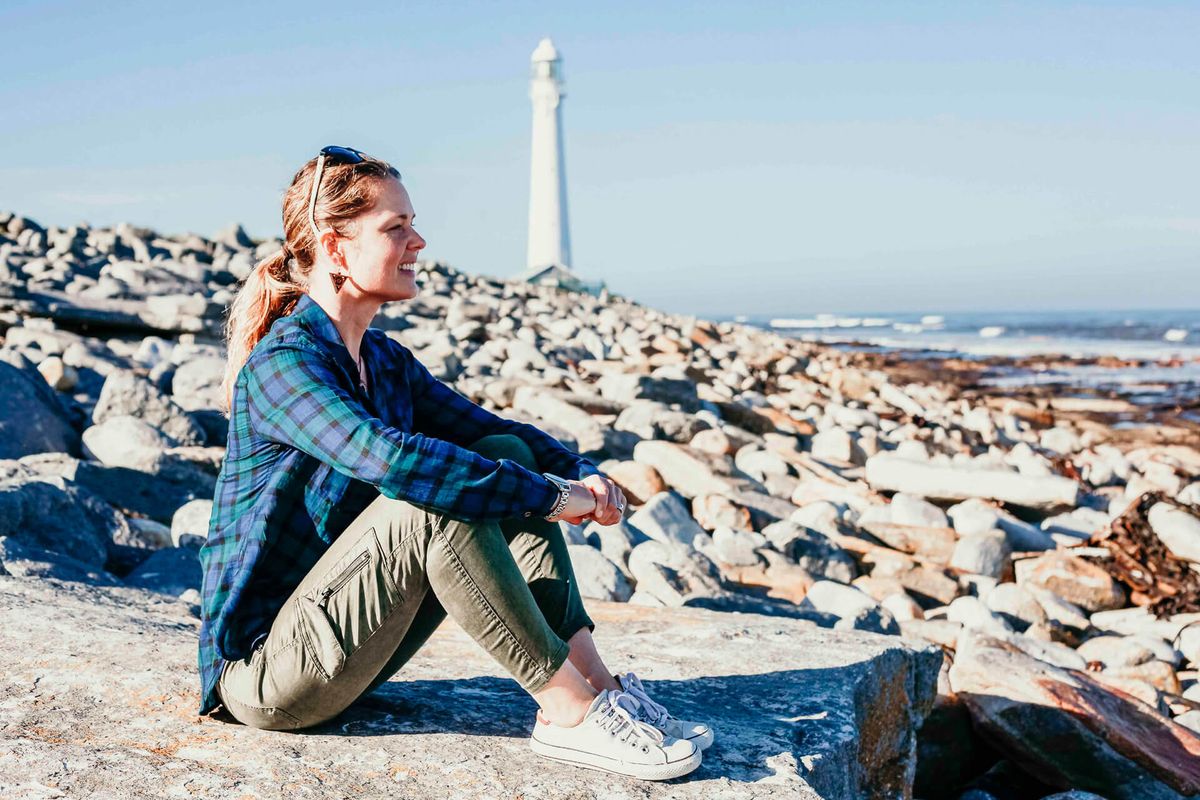 Nő ül egy sziklás tengerparton, világítótorony a háttérben