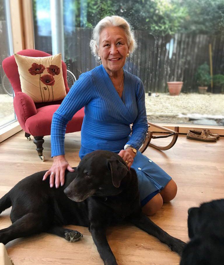 Ældre kvinde i en blå kjole i et beboelsesrum, knælende med en stor balckhund foran på sig